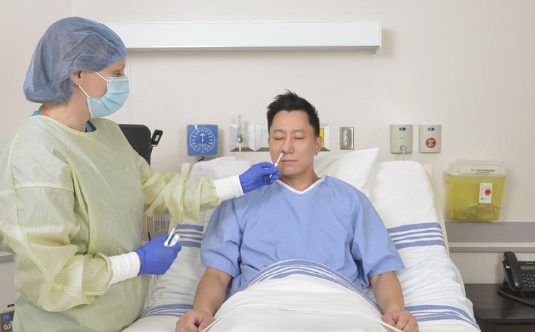 A female clinician in full PEE administers a nasal swab to a Asian male patient lysing in a hospital bed